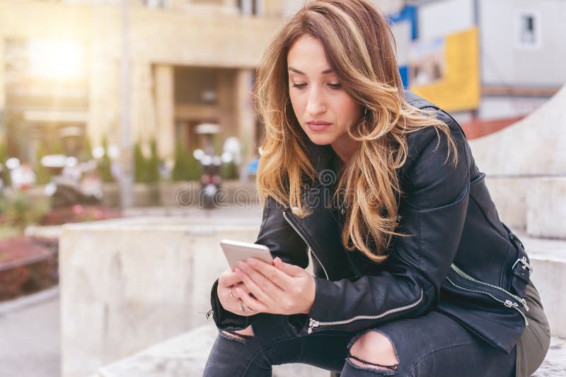 Mujer que se sienta en la calle de la ciudad, mensaje que mecanografía, charlando en smartp fotografía de archivo