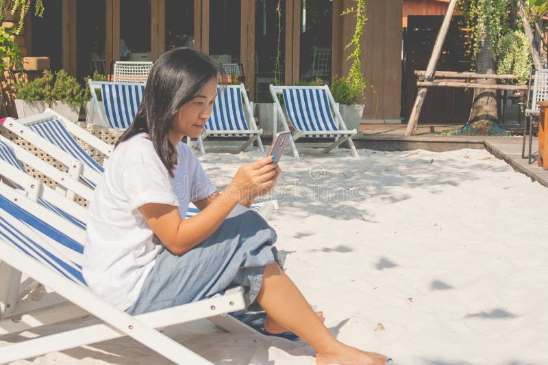 Mujer que se sienta en silla de playa y que juega smartphone en el exterior imagenes de archivo Mujer que se sienta en silla de playa y que juega smartphone en el exterior imagenes de archivo