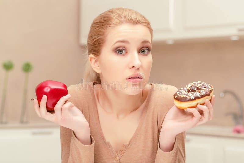 Being in doubt. Nice young blonde woman in her kitchen sits holding apple in one hand and donut in another hand hesitating and trying to choose what she has to eat. Being in doubt. Nice young blonde woman in her kitchen sits holding apple in one hand and donut in another hand hesitating and trying to choose what she has to eat