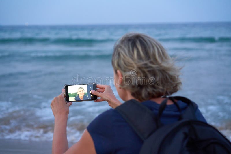 Mujer que toma a un autorretrato en el tel? fono elegante la playa en puesta del sol en fotografía de archivo libre de regalías