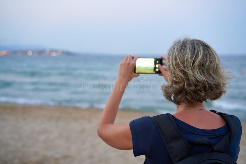 Mujer que toma una foto de un mar en el tel? fono elegante en puesta del sol fotos de archivo libres de regalías