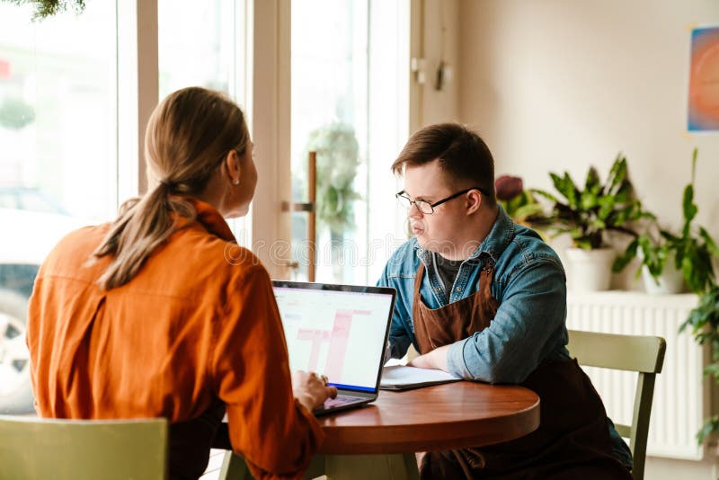 Mujer que usa una laptop mientras entrena a un hombre con síndrome de baja para trabajar en una cafetería foto de archivo libre de regalías