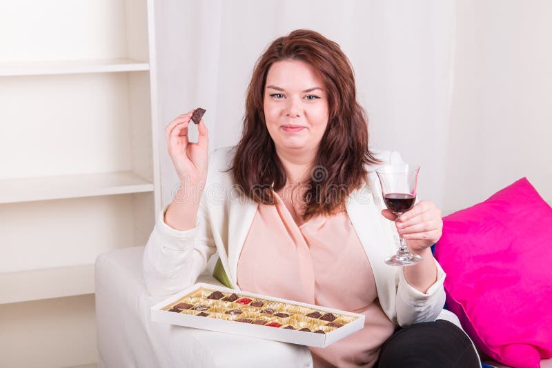 Mujer regordeta en casa que come los chocolates y que bebe el vino imagenes de archivo Mujer regordeta en casa que come los chocolates y que bebe el vino imagenes de archivo