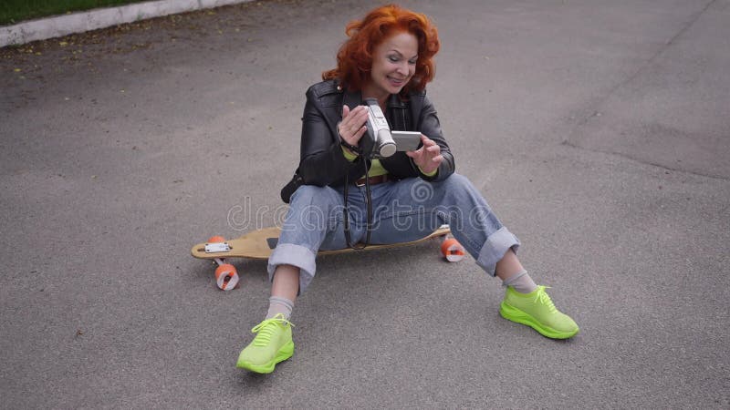 Mujer retro feliz de tiro ancho riendo sentada en skateboard viendo video en cámara vintage. retrato alegre almacen de metraje de vídeo