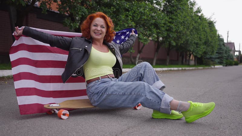 Mujer retro feliz sentada en patineta con bandera de los estados unidos mirando la cámara sonriendo. mujer caucásica pelirroja pos almacen de video