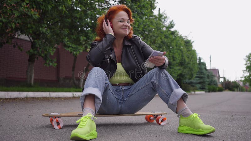 Mujer retro relajada y feliz sentada en skateboard encendiendo lista de reproducción escuchando música en auriculares. retrato de almacen de metraje de vídeo