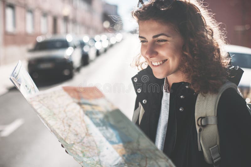 Mujer rizada sonriente con mochila mirando el mapa y viajando por la ciudad fotos de archivo libres de regalías