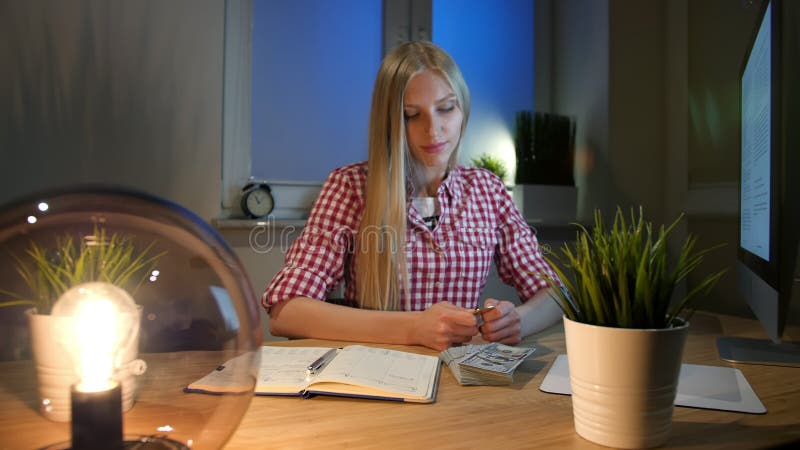 Mujer rubia en el escritorio que mira el bitcoin La sentada femenina rubia joven hermosa en el lugar de trabajo con el ordenador