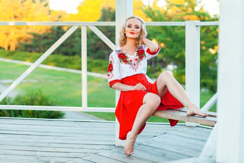 Mujer rubia joven magnífica con el maquillaje, peinado rizado en el vestido blanco rojo elegante que presenta en banco en cenador imagen de archivo Mujer rubia joven magnífica con el maquillaje, peinado rizado en el vestido blanco rojo elegante que presenta en banco en cenador imagen de archivo