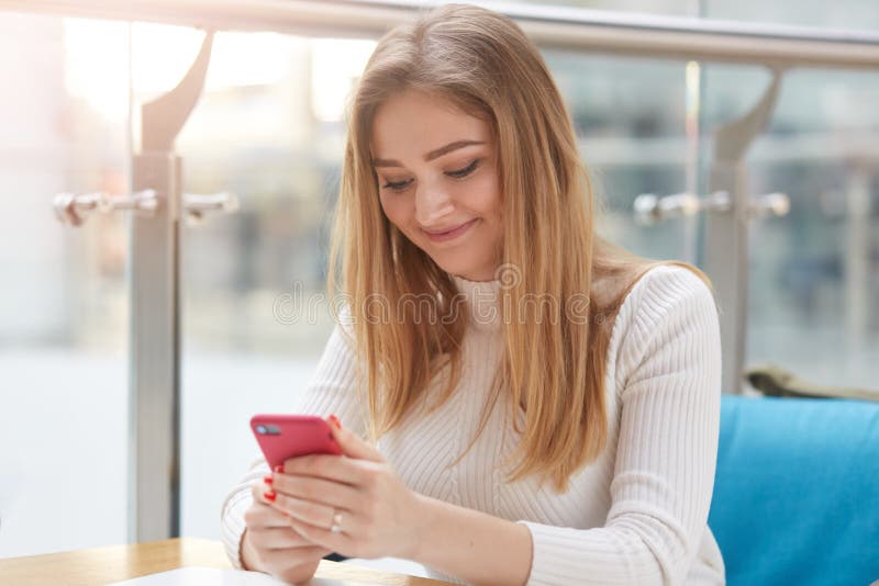 Mujer rubia joven sonriente con el pelo recto largo, en los masajes blancos de la camisa en su teléfono móvil mientras que se sie fotografía de archivo Mujer rubia joven sonriente con el pelo recto largo, en los masajes blancos de la camisa en su teléfono móvil mientras que se sie fotografía de archivo