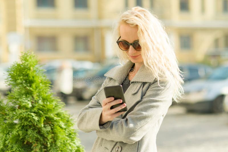 Mujer rubia sonriente adulta que habla en el teléfono celular, fondo de la calle de la ciudad, día soleado del otoño fotos de archivo Mujer rubia sonriente adulta que habla en el teléfono celular, fondo de la calle de la ciudad, día soleado del otoño fotos de archivo