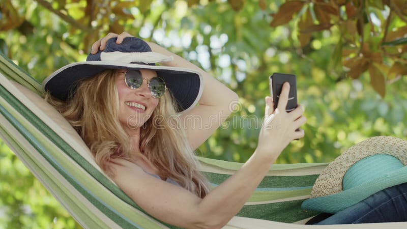 Mujer rubia sonriente con gafas de sol usando un smartphone cambia sus sombreros para hacer un video de desafío en la hamaca en el almacen de metraje de vídeo