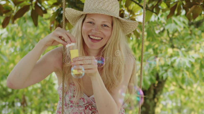 Mujer rubia sonriente con sombrero de paja hace burbujas de jabón sentada en el columpio en el jardín de casa ocio y vacaciones de almacen de metraje de vídeo