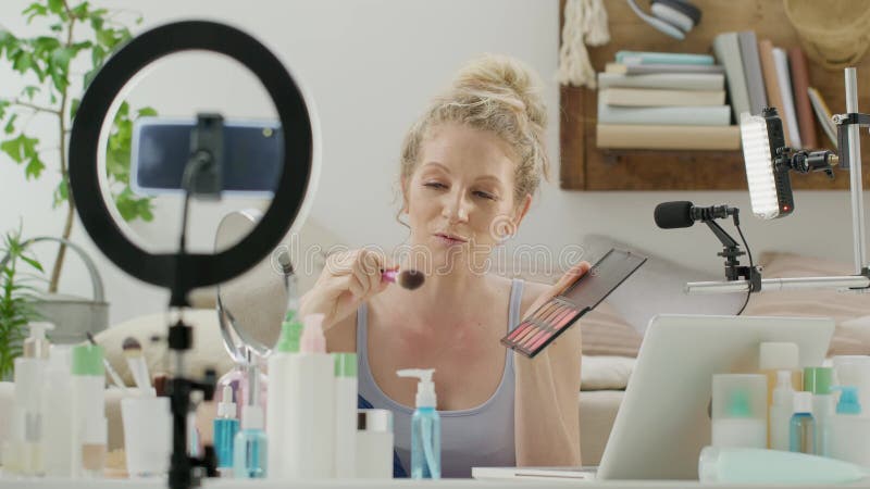 Mujer rubia sonriente influenciante blogger grabando un video tutorial en la cámara de un smartphone que muestra cómo aplicar cosm metrajes