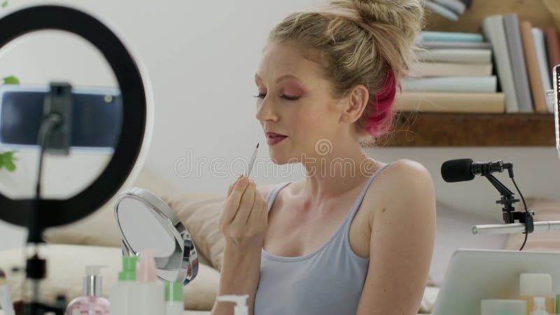 Mujer rubia sonriente influenciante blogger grabando un video tutorial en la cámara de un smartphone que muestra cómo aplicar cosm metrajes