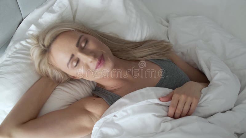 Mujer rubia sonriente que miente en su cama en dormitorio brillante debajo del edredón almacen de video