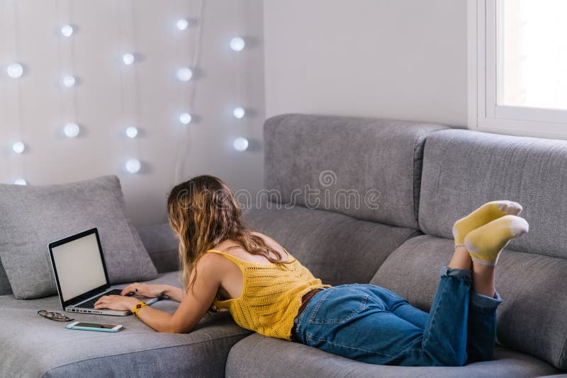Mujer rubia tendida en un sofá trabajando con un portátil en casa fotos de archivo