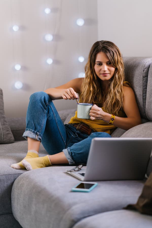 Mujer sentada en un sofá usando un portátil y tomando café. Feliz joven rubia sentada en un sofá usando un portátil para videollamadas y tomando café taza