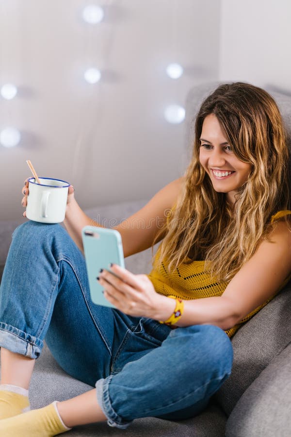 Mujer sentada en un sofá usando un teléfono inteligente y tomando café imagen de archivo