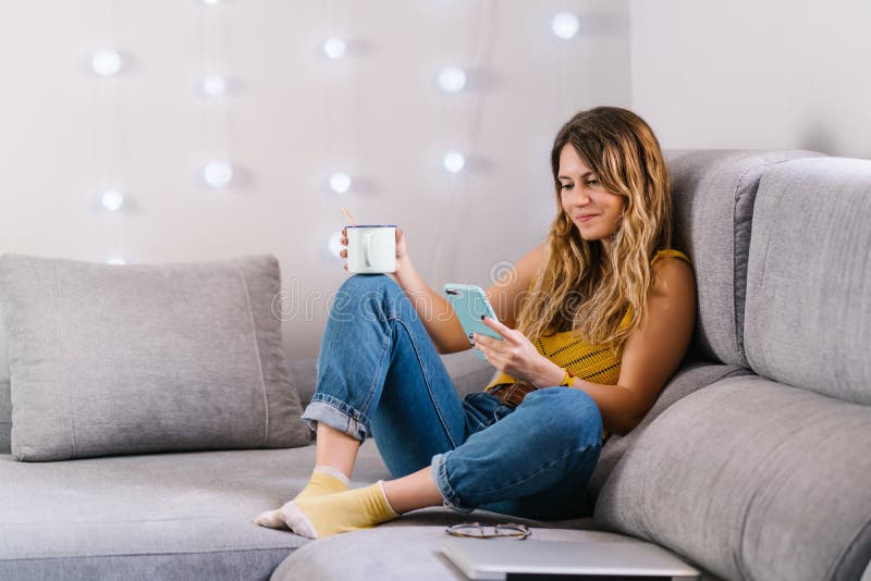 Mujer sentada en un sofá usando un teléfono inteligente y tomando café fotografía de archivo libre de regalías