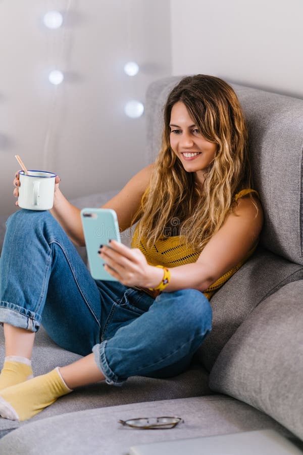 Mujer sentada en un sofá usando un teléfono inteligente y tomando café imagen de archivo libre de regalías