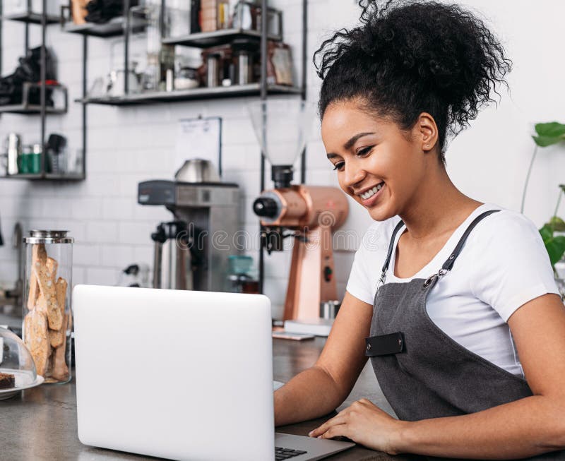 Mujer sonriente con el pelo rizado usando un delantal tecleando en una laptop imagenes de archivo