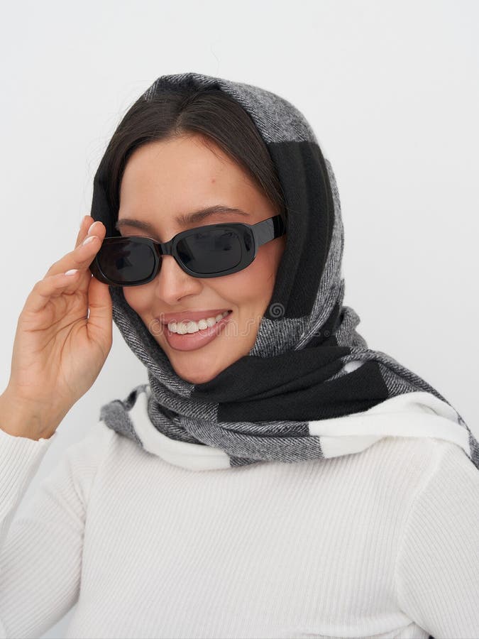 Mujer sonriente con gafas de sol rectangulares y una bufanda de cuadros negros y grises sobre su cabeza. Su mano se ajusta fotografía de archivo