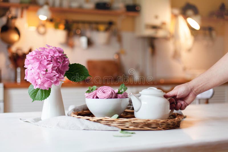 Mujer toma tetera blanca de servir bandeja con malvaviscos rosas. La flor de Hydrangea está en la mesa de la cocina fotos de archivo libres de regalías Mujer toma tetera blanca de servir bandeja con malvaviscos rosas. La flor de Hydrangea está en la mesa de la cocina fotos de archivo libres de regalías