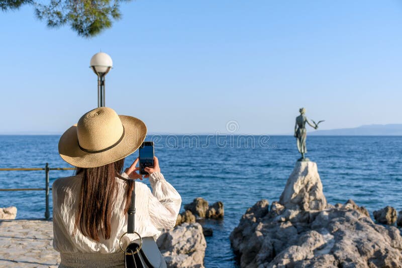 Mujer tomando fotos de una estatua de una doncella con gaviota en la costa rocosa en opatija croacia. foto de archivo
