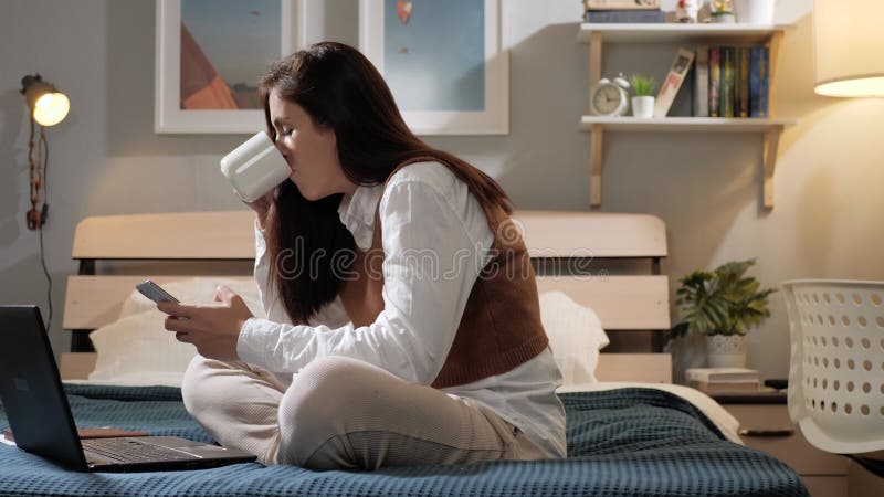 Mujer usa teléfono y bebe café. mujer sentada en la cama en el dormitorio y escribe texto en la pantalla de un smartphone y bebe c metrajes