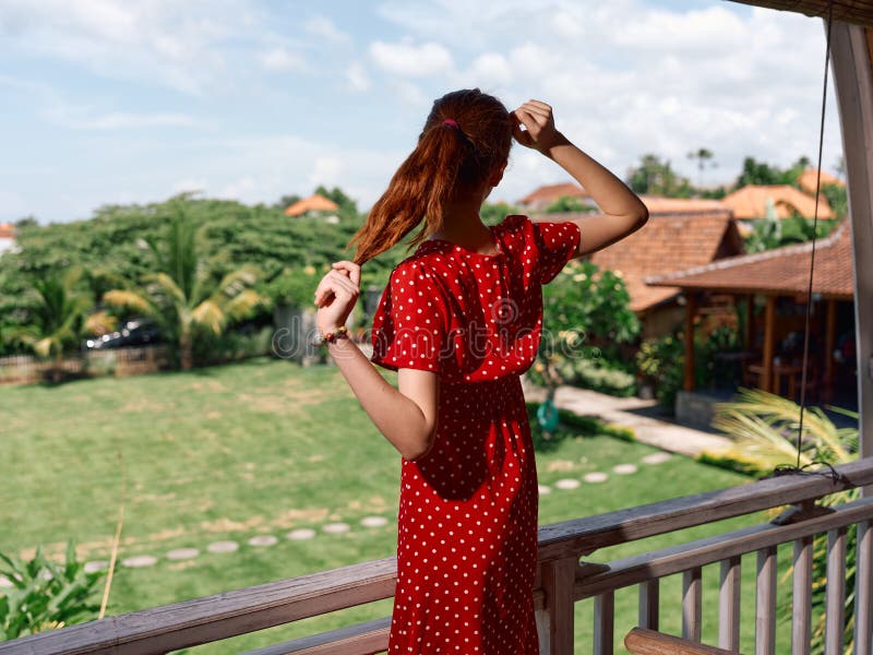 Mujer vestida con un punto polka rojo admirando el exuberante césped verde desde el balcón en los días soleados imagen de archivo