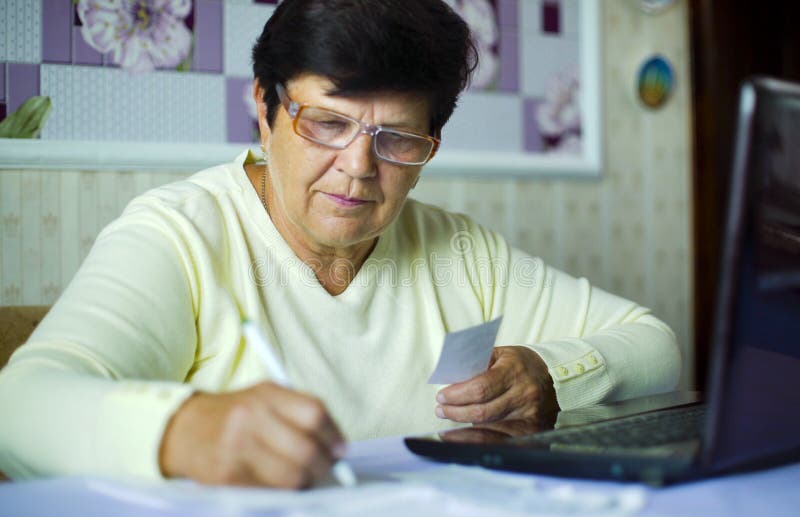 Mulher adulta superior nos monóculos que verifica custos de despesas diárias no portátil em casa fotografia de stock Mulher adulta superior nos monóculos que verifica custos de despesas diárias no portátil em casa fotografia de stock