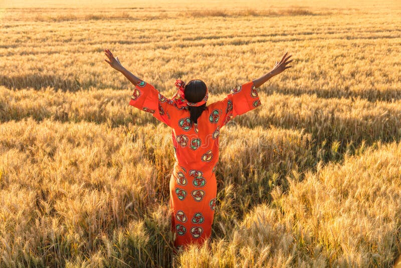 African woman in traditional clothes standing arms raised in a field of barley or wheat crops at sunset or sunrise. African woman in traditional clothes standing arms raised in a field of barley or wheat crops at sunset or sunrise