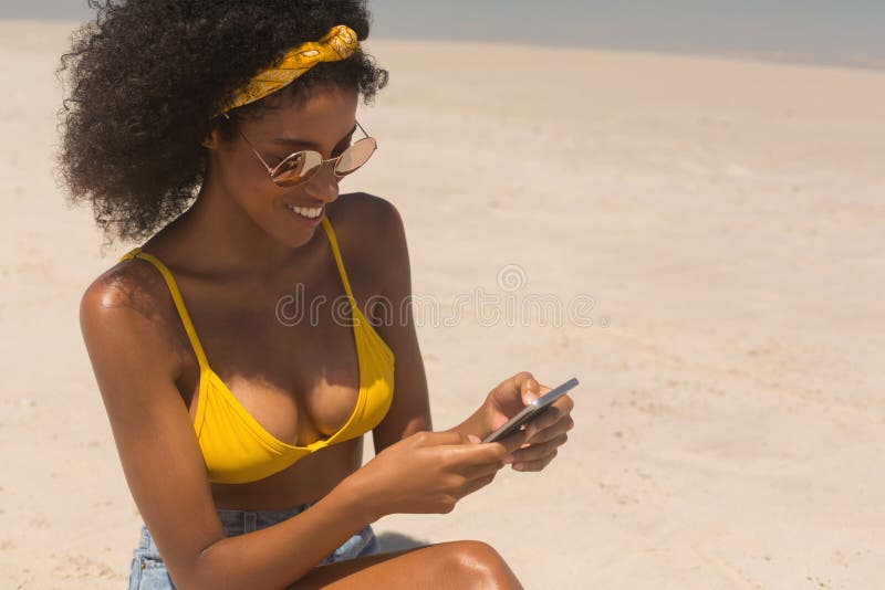 Mulher afro-americano nova no biquini e em óculos de sol amarelos usando o telefone celular na praia imagem de stock royalty free Mulher afro-americano nova no biquini e em óculos de sol amarelos usando o telefone celular na praia imagem de stock royalty free