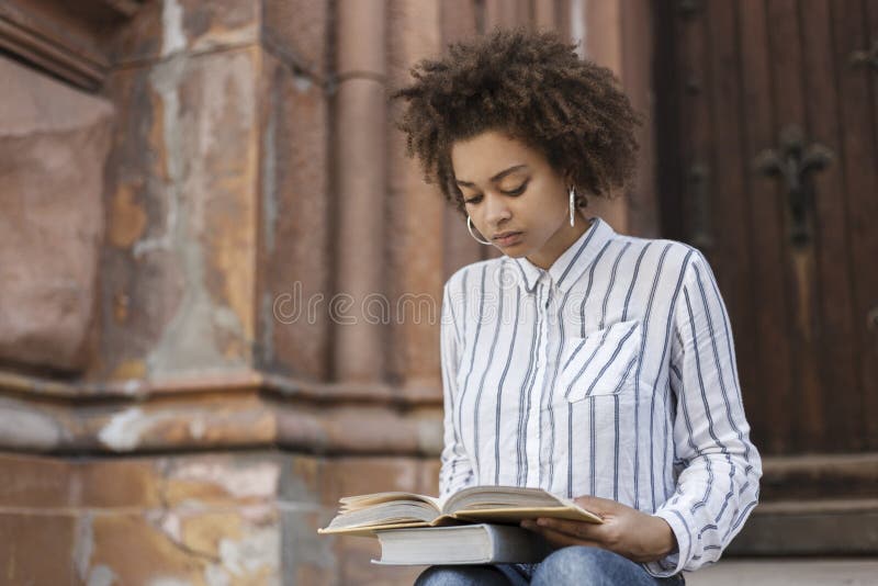 Mulher afro-americano que senta-se na rua e que lê um livro Em livros dos pés Há uma construção ao lado de uma menina de pele esc imagem de stock Mulher afro-americano que senta-se na rua e que lê um livro Em livros dos pés Há uma construção ao lado de uma menina de pele esc imagem de stock