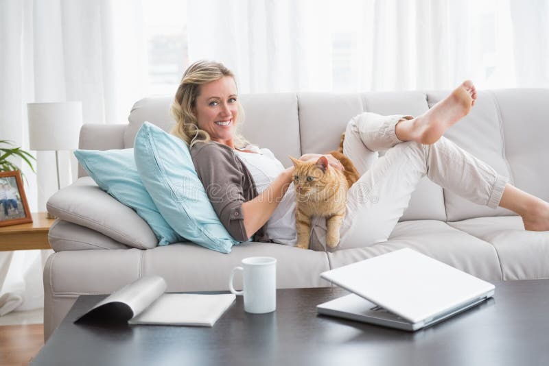 Mulher alegre que encontra-se no sofá que afaga um gato do gengibre imagens de stock Mulher alegre que encontra-se no sofá que afaga um gato do gengibre imagens de stock