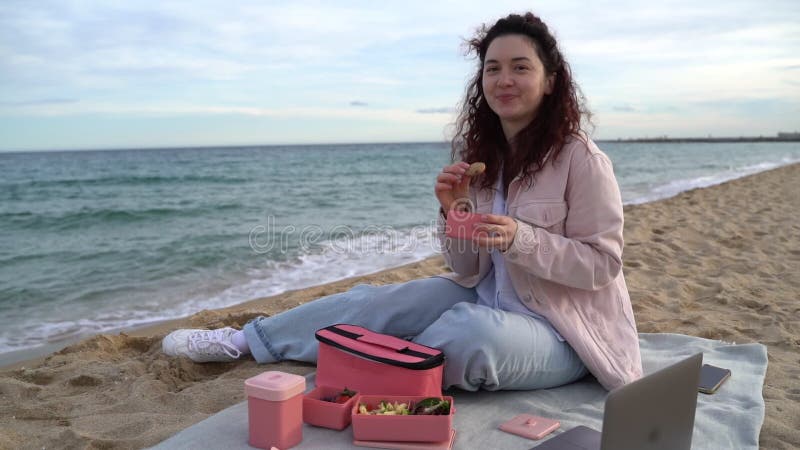 Mulher almoçando na praia comendo biscoitos de chocolate de plástico rosa.