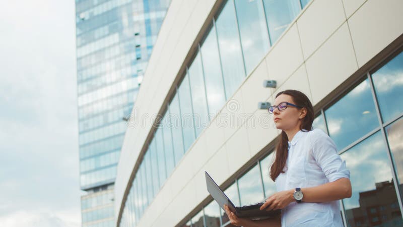 Mulher ambiciosa nova com um portátil em suas mãos no fundo do centro de negócios imagem de stock Mulher ambiciosa nova com um portátil em suas mãos no fundo do centro de negócios imagem de stock