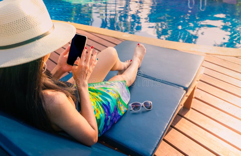 A mulher asiática com chapéu e o roupa de banho sentam-se no sunbed na piscina e smartphone da utilização em férias de verão pela foto de stock royalty free A mulher asiática com chapéu e o roupa de banho sentam-se no sunbed na piscina e smartphone da utilização em férias de verão pela foto de stock royalty free