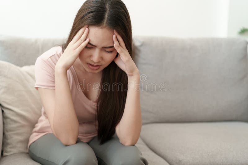 A mulher asiática está guardando suas mãos à cabeça na dor no sofá em casa, jovens mulheres tem dores de cabeça severas das enxaq fotografia de stock A mulher asiática está guardando suas mãos à cabeça na dor no sofá em casa, jovens mulheres tem dores de cabeça severas das enxaq fotografia de stock