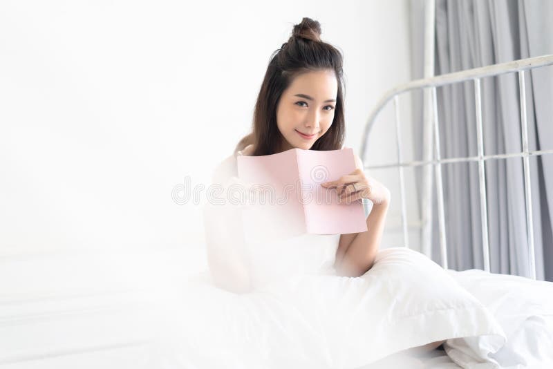 Mulher asiática nova bonita bonito que lê um livro e que sorri como se senta na cama, olhando a câmera Estilo de vida da mulher d imagem de stock royalty free Mulher asiática nova bonita bonito que lê um livro e que sorri como se senta na cama, olhando a câmera Estilo de vida da mulher d imagem de stock royalty free