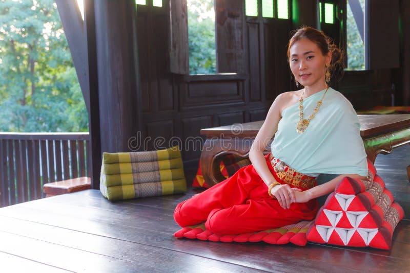 A mulher asiática tailandesa bonita nova que veste o traje tailandês tradicional do vintage senta-se no coxim do triângulo no bal foto de stock A mulher asiática tailandesa bonita nova que veste o traje tailandês tradicional do vintage senta-se no coxim do triângulo no bal foto de stock