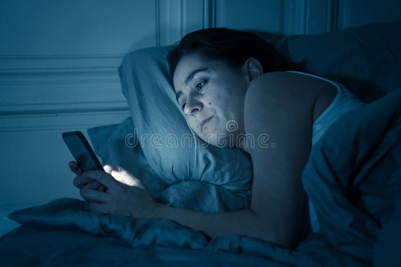 Mulher atrativa do latino dedicada ao telefone celular e ao Internet tarde em nigh na cama que olha sem sono fotografia de stock Mulher atrativa do latino dedicada ao telefone celular e ao Internet tarde em nigh na cama que olha sem sono fotografia de stock
