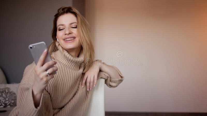 A mulher atrativa nova em uma camiseta senta-se em casa na sala de visitas e usa-se um smartphone foto de stock A mulher atrativa nova em uma camiseta senta-se em casa na sala de visitas e usa-se um smartphone foto de stock