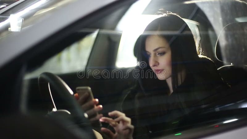 Mulher atrativa nova que usa o telefone celular no carro no estacionamento subterrâneo Movimento lento