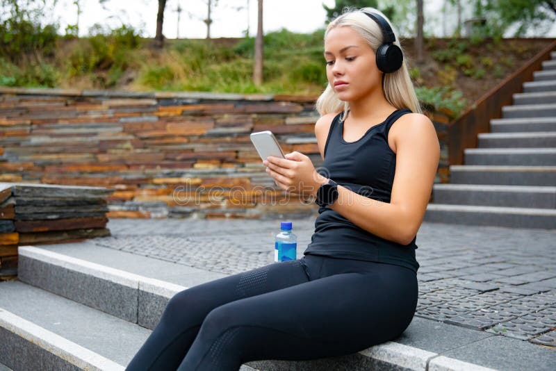 A mulher bonita dos esportes que senta-se em escadas escuta a música do smartphone imagem de stock royalty free A mulher bonita dos esportes que senta-se em escadas escuta a música do smartphone imagem de stock royalty free