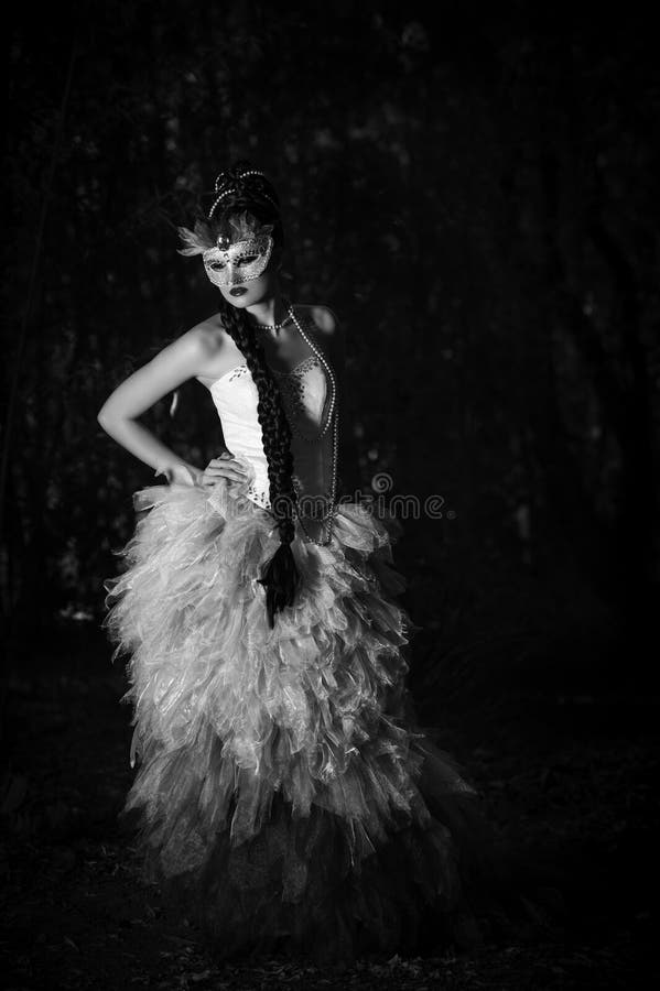 Monochrome image of a beautiful young Caucasian woman wearing her long dark hair in an elaborate braided hairstyle with beaded necklaces, wearing a couture evening dress and an ornate mask, while she stands in a forest. Monochrome image of a beautiful young Caucasian woman wearing her long dark hair in an elaborate braided hairstyle with beaded necklaces, wearing a couture evening dress and an ornate mask, while she stands in a forest