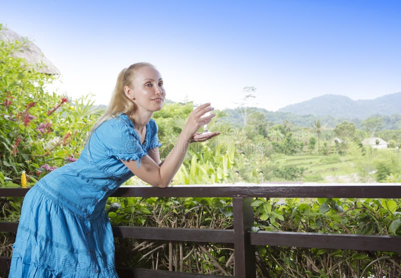 A mulher bonita nova em um vestido longo com um vidro do vinho olha a natureza tropical fotos de stock A mulher bonita nova em um vestido longo com um vidro do vinho olha a natureza tropical fotos de stock