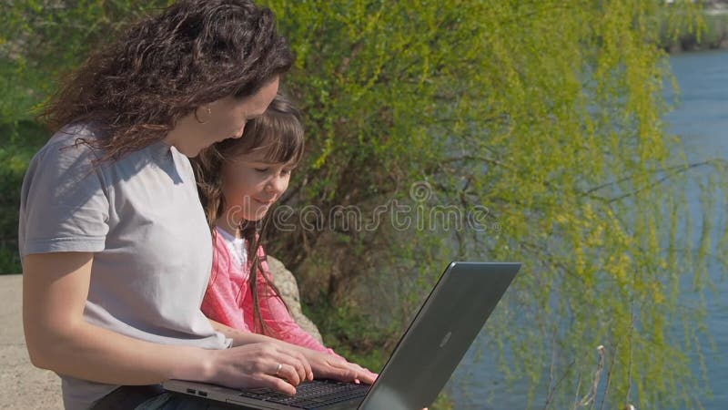 Mulher com bebê e portátil fora Família feliz no banco de rio A mãe está ensinando uma criança em um portátil Um dia ensolarado d