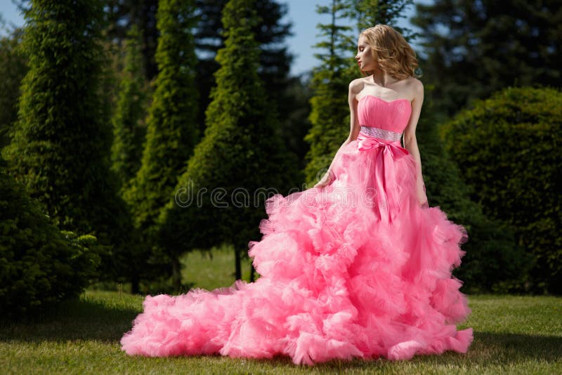 A mulher com os fechamentos louros que vestem o vestido de noite cor-de-rosa com saia macia está levantando no jardim botânico na imagem de stock A mulher com os fechamentos louros que vestem o vestido de noite cor-de-rosa com saia macia está levantando no jardim botânico na imagem de stock